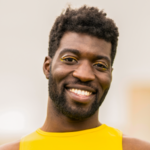 A Black person with a short beard and short hair smiling at the camera, wearing a yellow tank top and dark yellow eye shadow.