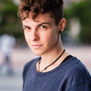 A white androgynous person with short dark brown hair, wearing a navy blue shirt, smirking and looking at the camera.
